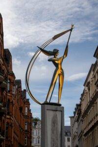 Eine moderne gold-schwarze Skulptur einer Figur mit Bogen, die als Symbol für die lebendigen Veranstaltungen in Flensburg steht, ragt vor einer Reihe traditioneller roter Backsteingebäude in die Höhe, und verbindet die reiche Geschichte der Stadt mit ihrem dynamischen kulturellen Angebot
