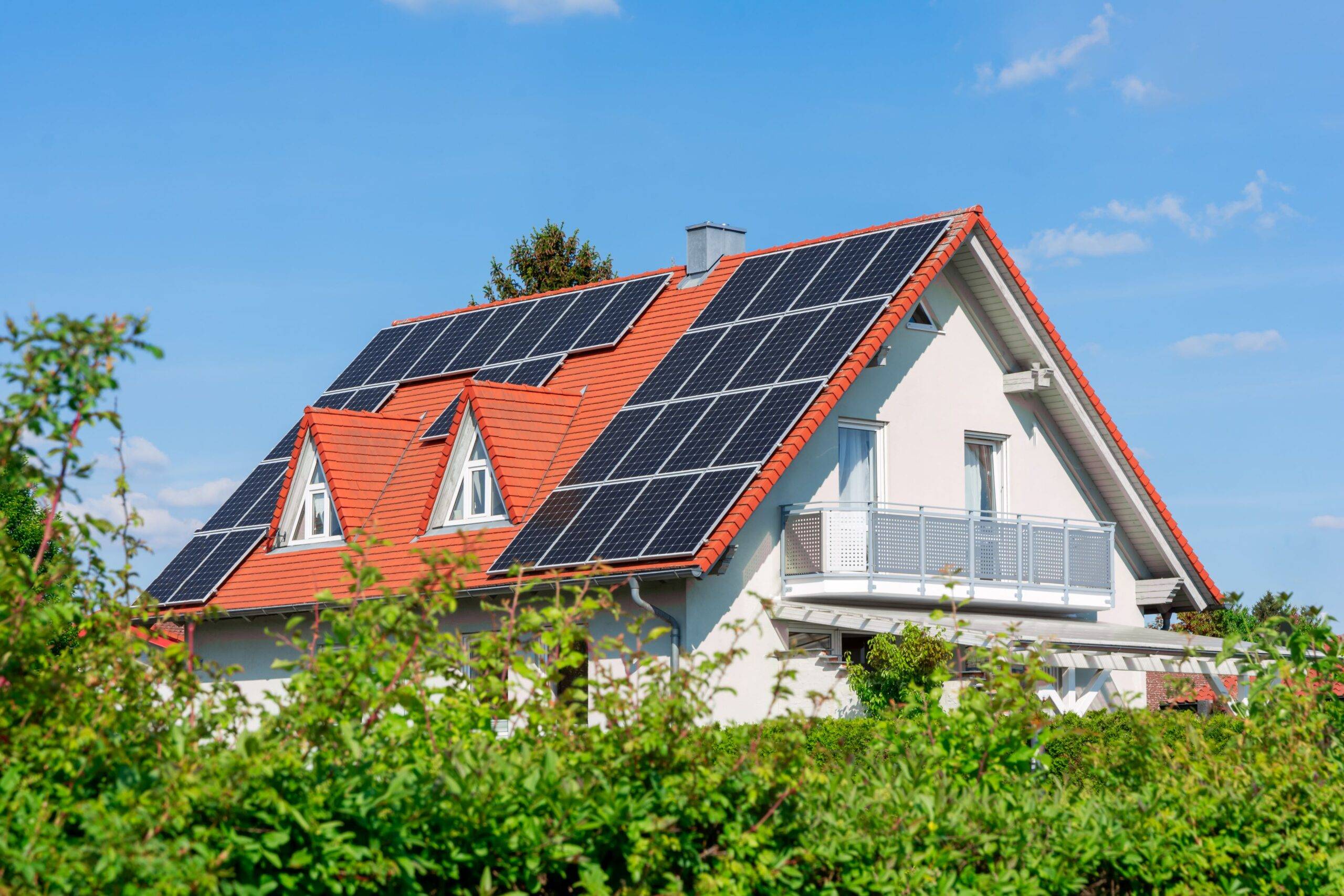 Ein Einfamilienhaus mit rotem Ziegeldach, auf dem mehrere Solarmodule installiert sind. Das Haus steht an einem sonnigen Tag unter einem blauen Himmel, umgeben von grünen Büschen.