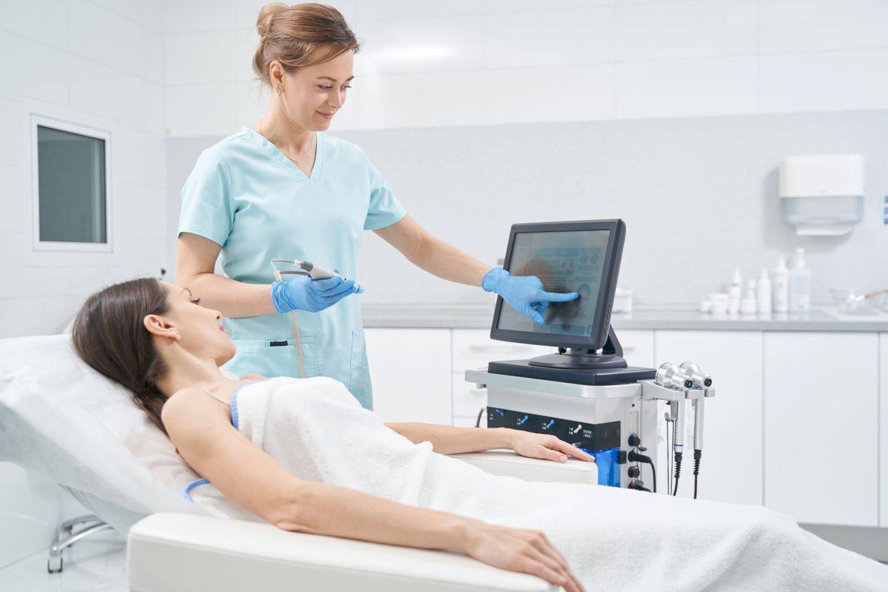 Kosmetikerin erklärt der Kundin die Hydrafacial Behandlung, während sie das Gerät für die Tiefenreinigung und Pflege einstellt