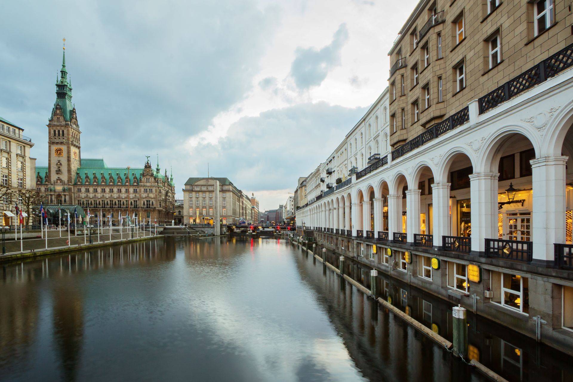 Blick auf die Alsterarkaden und das Hamburger Rathaus bei ruhigem Wasser und bewölktem Himmel