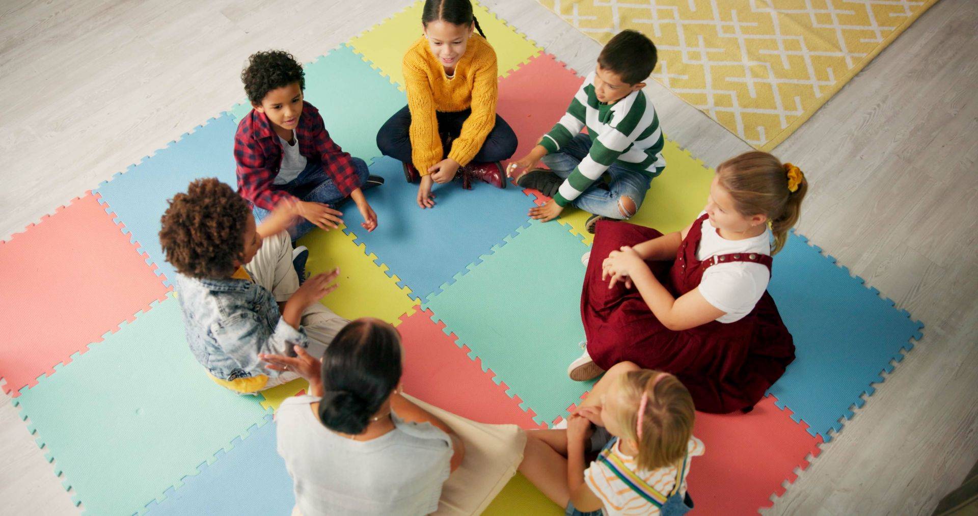Mehrere Kinder sitzen im Kreis auf bunten Spielmatten und sprechen mit einer pädagogischen Fachkraft – typischer Moment im Kita-Alltag